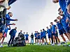 Fotballspillere og trenere samles i en ring på gressbanen under trening.