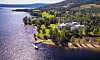 Utsikt over en kyststripe med skog, åpent landskap, brygge og bygninger langs vannet.