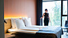 Lyst hotellrom med stor seng, nattbord, varme lamper og en person ved gulv-til-tak-vinduene.