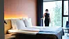 Lyst hotellrom med stor seng, nattbord, varme lamper og en person ved gulv-til-tak-vinduene.
