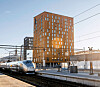 Høyhastighetstog ved en moderne stasjon med høy, brun bygning og jernbanespor.