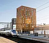 Høyhastighetstog ved en moderne stasjon med høy, brun bygning og jernbanespor.