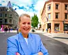 Smilende kvinne i blå blazer står foran bygninger i en solfylt gate.