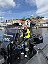 Båtfører i gul og svart dress står på en svart RIB-båt ved en småbåthavn.