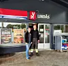To ansatte står foran inngangen til en Lindås-butikk med rødt og hvitt skilt.