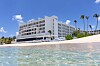 Hvitt hotell ved en sandstrand med palmer og klart vann under blå himmel.
