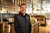 Smilende ansatt i svart vest står i et moderne restaurantlokale med bar og sitteplasser.