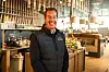 Smilende ansatt i svart vest står i et moderne restaurantlokale med bar og sitteplasser.