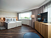 Lyst hotellrom med seng, store vinduer, gardiner, TV og trepanel på møbelveggen.
