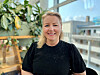 Smilende kvinne i svart topp foran store vinduer og planter i et lyst kontor.