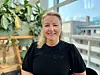 Smilende kvinne i svart topp foran store vinduer og planter i et lyst kontor.