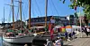 Seilbåter fortøyd ved en brygge med mennesker, markedsboder og bygninger langs havnen.