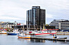 Fiskebåter i en havn foran et høyt mørkt bygg og bybebyggelse.