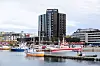 Fiskebåter i en havn foran et høyt mørkt bygg og bybebyggelse.