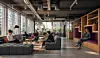 Moderne kontorlounge med mennesker, arbeidsplasser, planter og store vinduer.