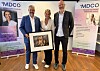 Fire personer holder et innrammet bilde foran MDCO-bannere i et lyst lokale.