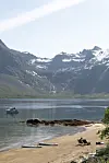 Innsjø med snødekte fjell, liten båt og person på en sandstrand.