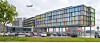 Moderne glassbygning ved en vei, med busser, trær og et fly på himmelen.