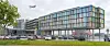 Moderne glassbygning ved en vei, med busser, trær og et fly på himmelen.