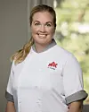 Smilende kokk i hvit uniform står foran et vindu med grønn bakgrunn.