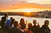 Silhuetter av mennesker ser en oransje solnedgang over vannet og byen.