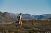 Vandrer med ryggsekk i lynglandskap foran fjell og blå himmel.