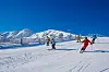 Skiløpere i en solfylt alpinbakke med snødekte fjell i bakgrunnen.