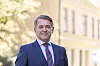Dresskledd mann foran en lys bygning og grønne trær.