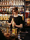 Bartender måler opp ingredienser bak en bar med flasker, glass og drinkutstyr.
