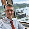 Mann i skjorte og slips ved et vindu med utsikt over vann, bygninger og fjell.