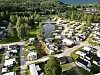 Luftfoto av campingplass med bobiler, campingvogner og hytter ved en skogkledd innsjø.