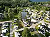 Luftfoto av campingplass med bobiler, campingvogner og hytter ved en skogkledd innsjø.