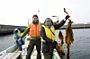 Tre personer i arbeidsklær står i en liten båt og viser fram brune vannplanter.