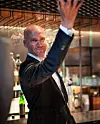 Mann i mørk dress hilser i en elegant bar med hånden løftet.