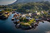 Luftfoto av en kystlandsby med hus og båter ved en fjord, omgitt av grønne fjell.