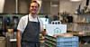 Kjøkkenarbeider i mørkt forkle står ved plastkasser og en papirpose på et storkjøkken.