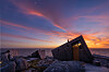 Trekantet trehytte på svaberg ved havet i en fargerik solnedgang.