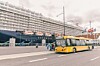 Gul bybuss kjører foran et stort cruiseskip ved en havn.