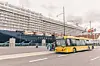 Gul bybuss kjører foran et stort cruiseskip ved en havn.