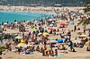 Fullsatt sandstrand med badende, fargerike parasoller og turkist vann.