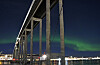 Grønt nordlys over Tromsøbrua om natten, med opplyste bygninger og speilinger i vannet.