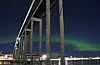 Grønt nordlys over Tromsøbrua om natten, med opplyste bygninger og speilinger i vannet.